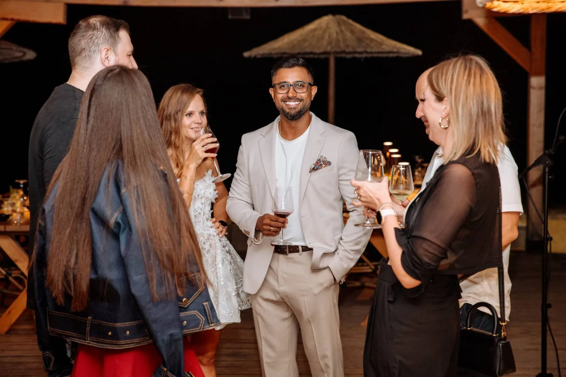 Dhee in a cream suit with a glass of red wine, laughing with friends at an evening party.
