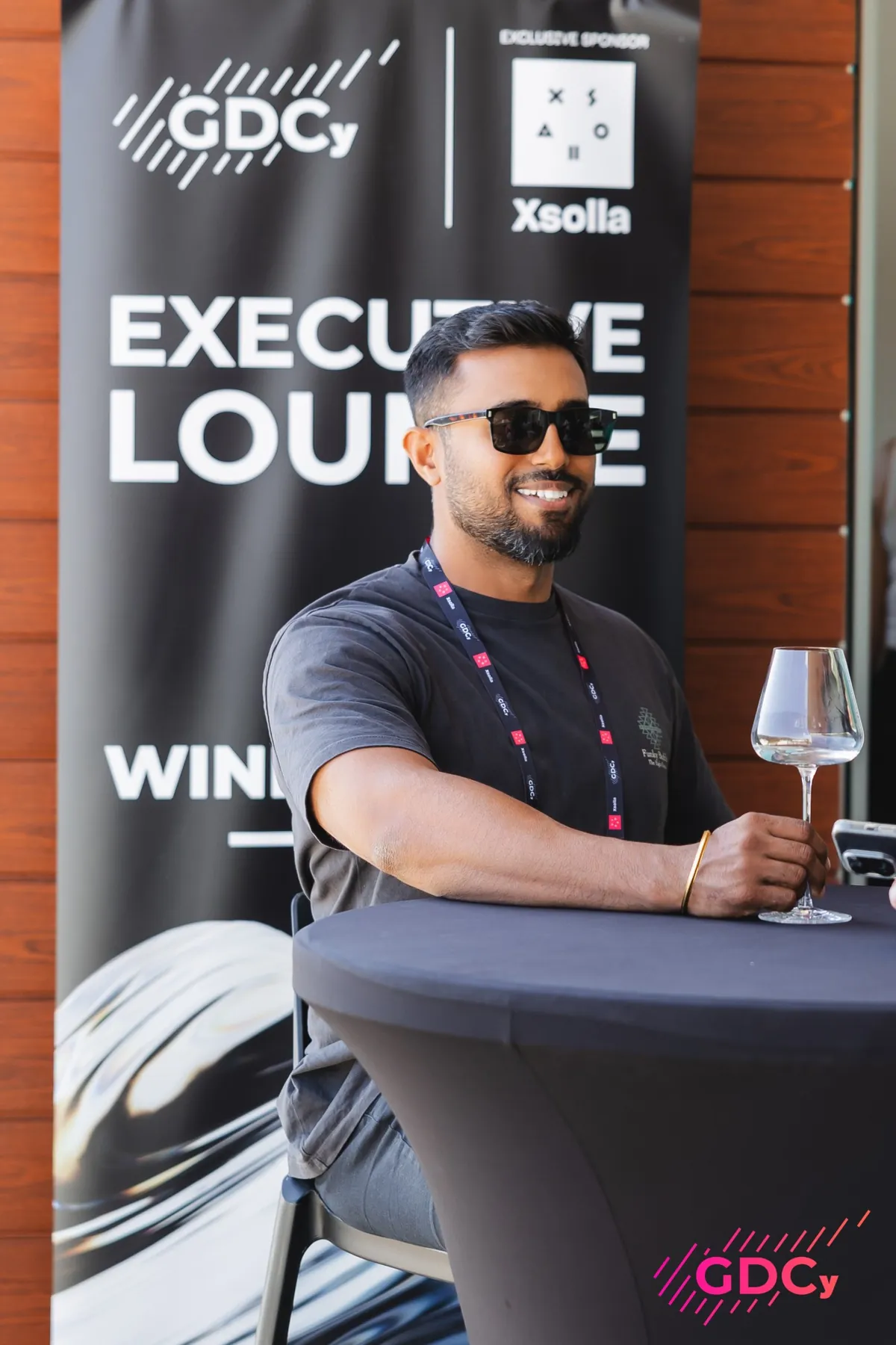Dhee in sunglasses with a glass of wine in a conference executive lounge.