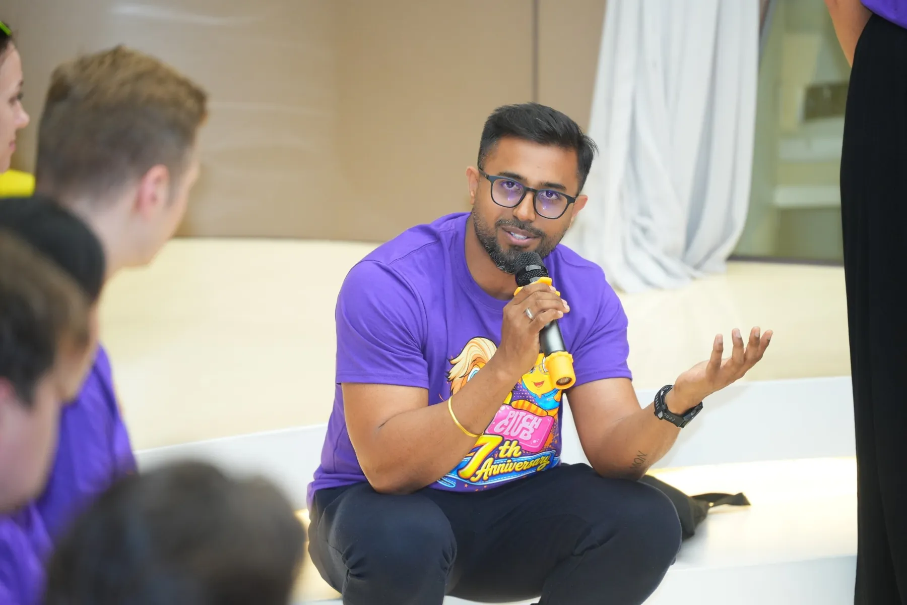 Dhee crouched with a microphone, mid-question, gesturing with an open hand.