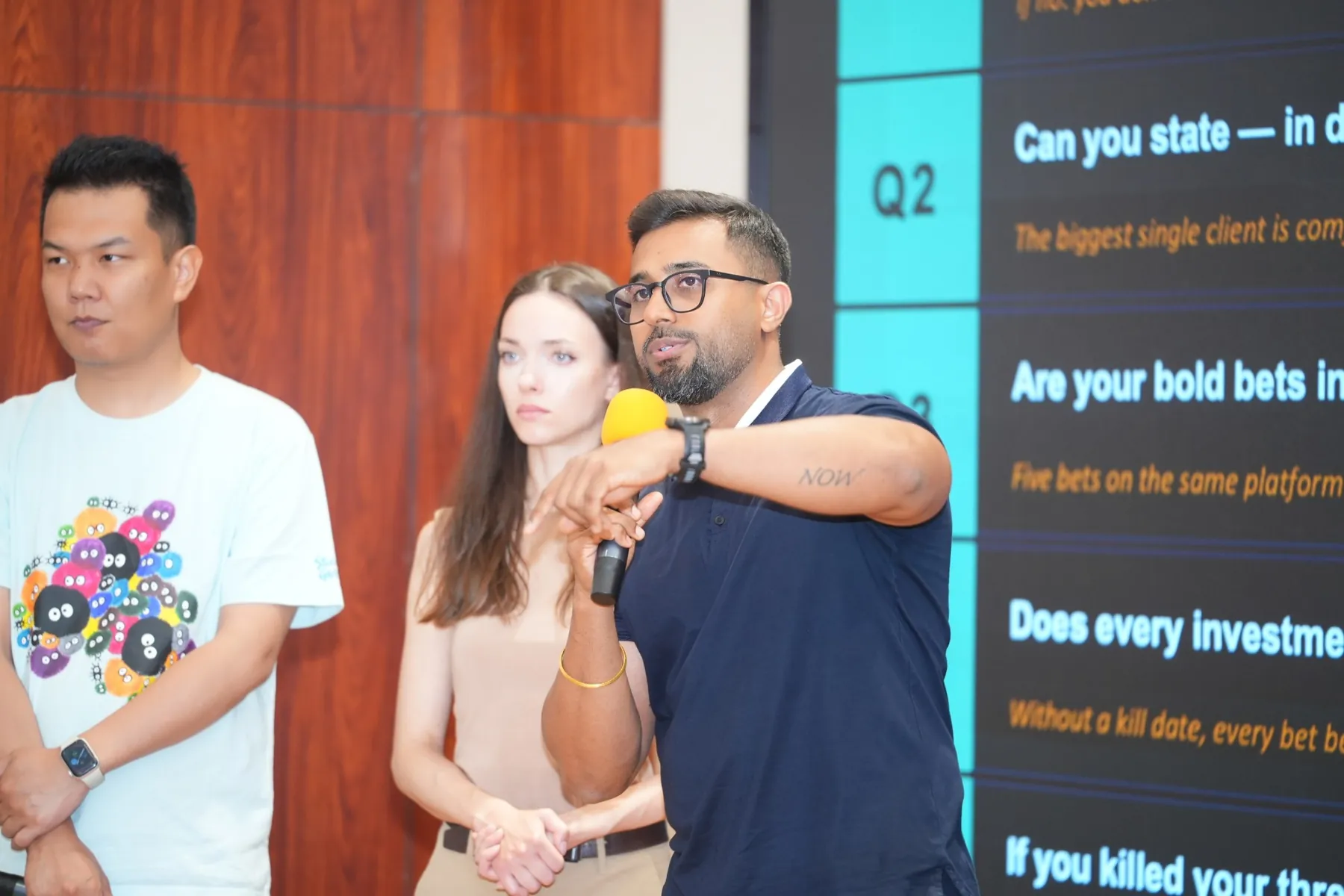 Dhee speaking into a microphone; the word NOW is tattooed on his forearm.